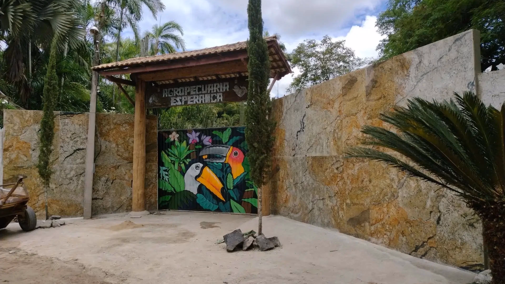 Entrada de fazenda imponente com muro de pedras naturais e portão de madeira, destacando a pintura artística de tucanos